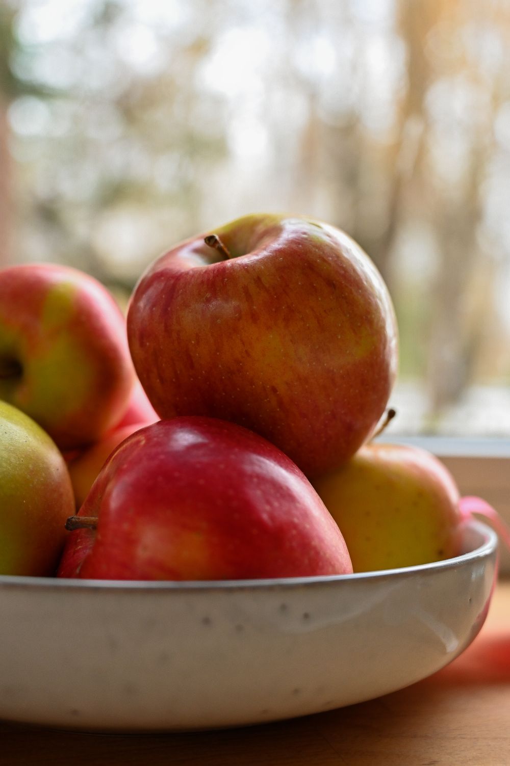 Whole red and yellow apples in a white ceramic bowl beside a window in natural light