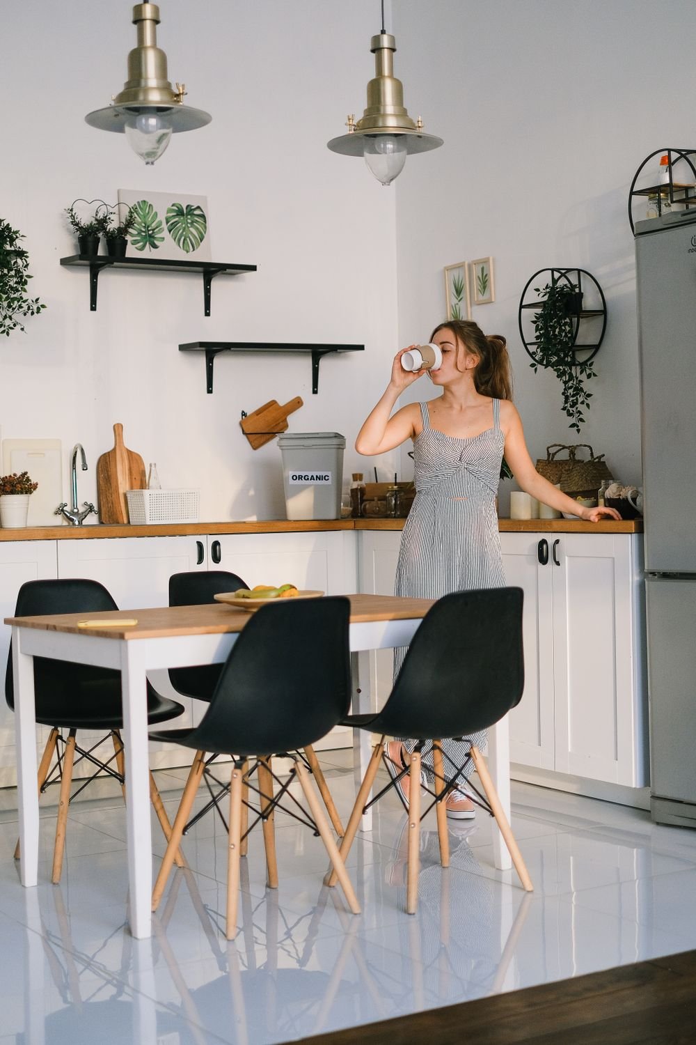 A woman standing in a bright modern kitchen drinking a cup of coffee, with an empty table and no food in sight