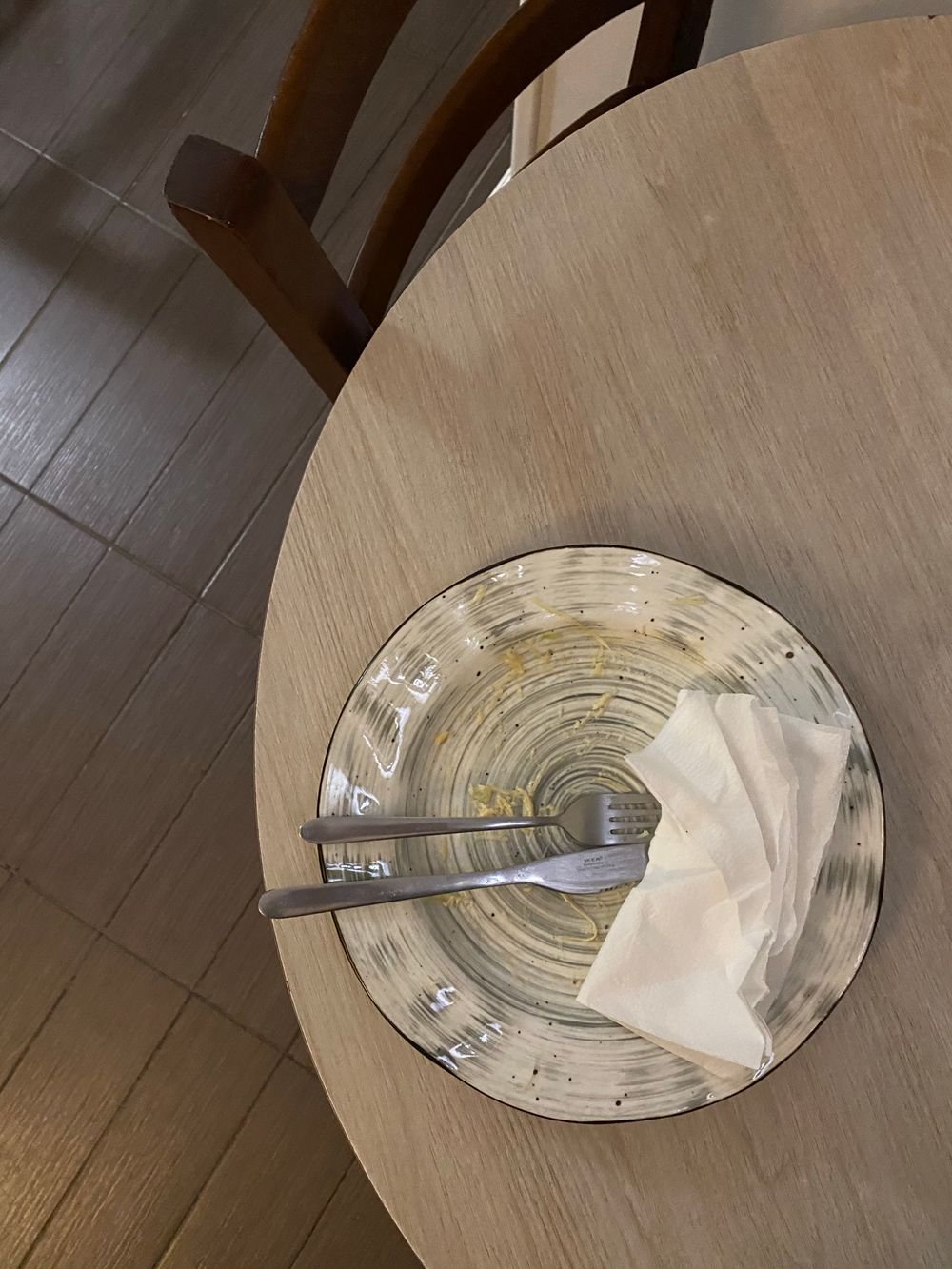 An empty plate with a fork and knife resting on a round wooden table after a finished meal