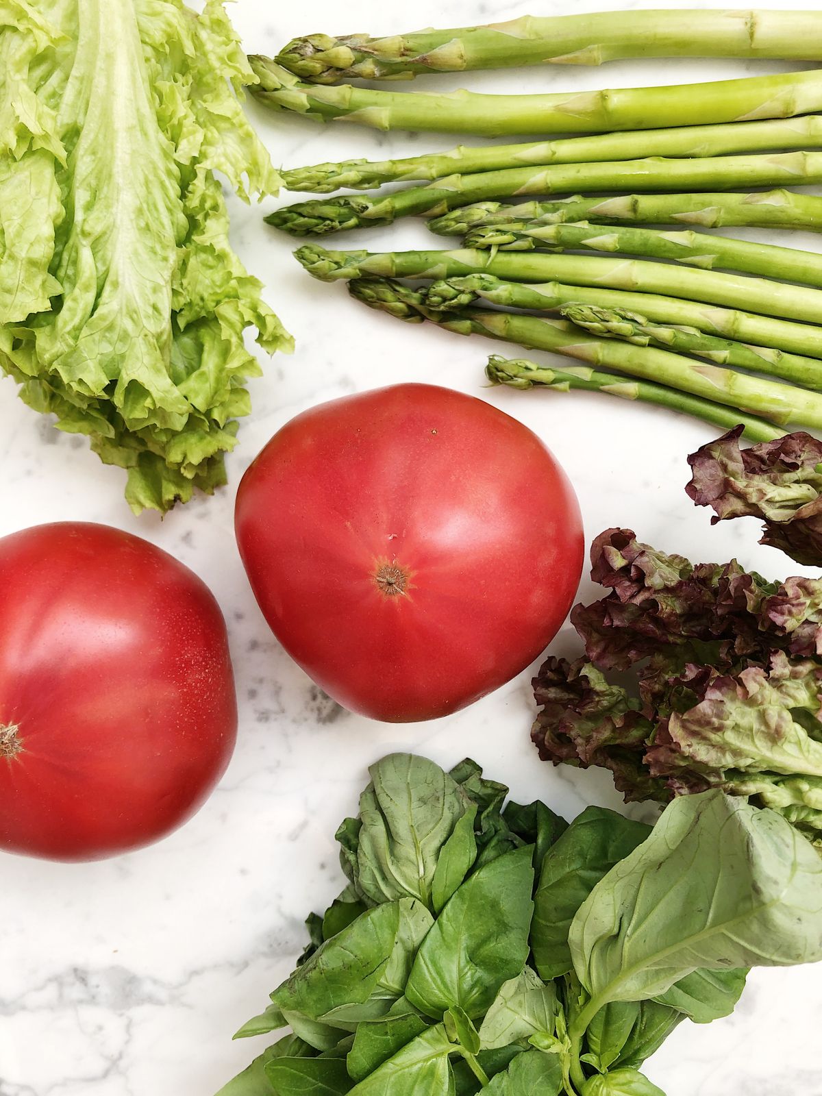 Flat lay of fresh asparagus, lettuce, basil and tomatoes on a marble surface, representing high-fiber whole foods
