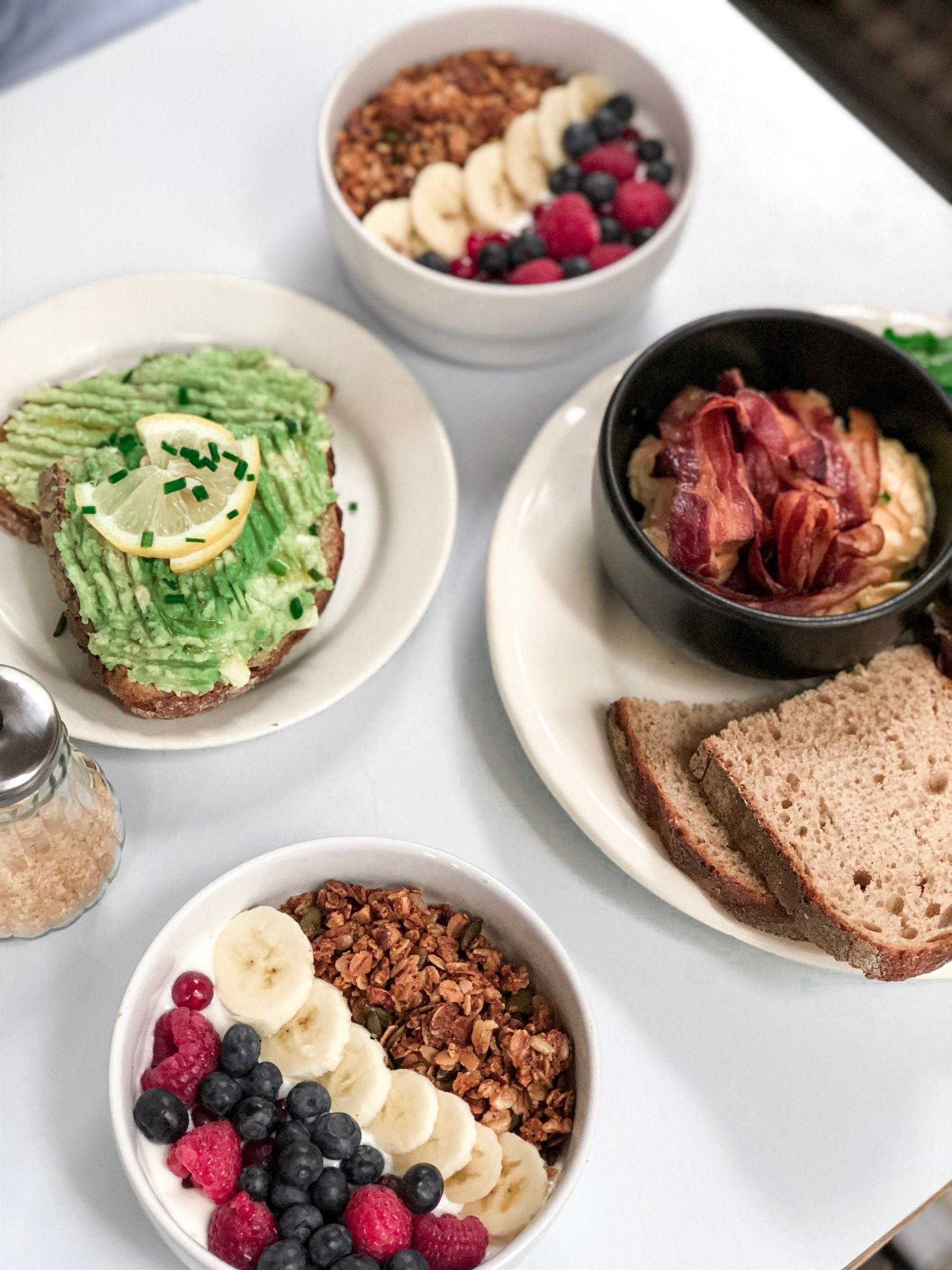 An overhead breakfast table with avocado toast, yogurt bowls topped with berries, granola and banana, and whole-grain bread