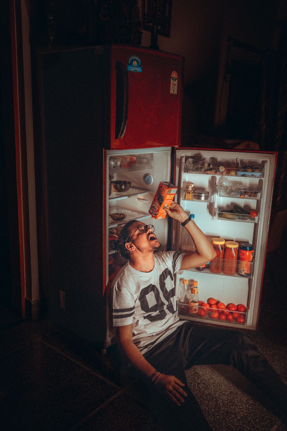 A person standing at an open refrigerator late at night, lit only by the fridge light, reaching for a snack