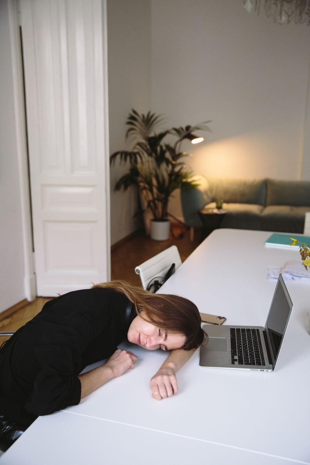 Woman resting her head on a white desk beside an open laptop in a bright home office, eyes closed after lunch