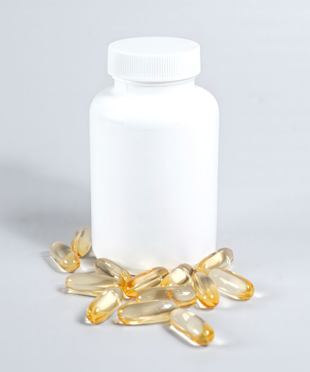 A plain white supplement bottle with no label, surrounded by golden softgel capsules on a white surface