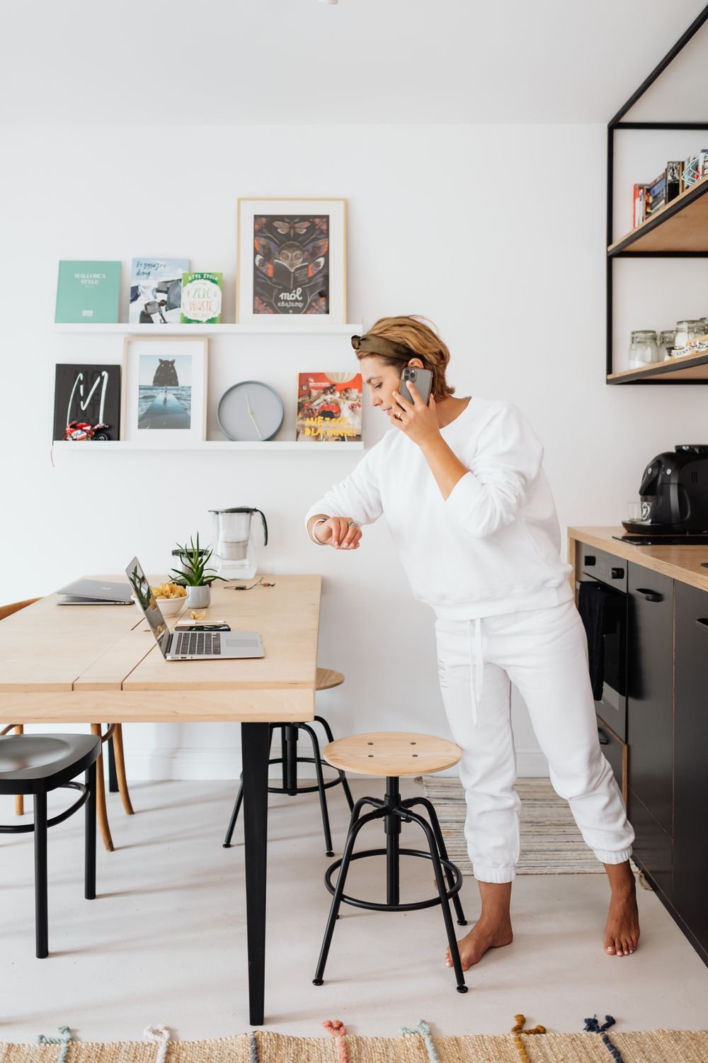 A woman in a bright modern kitchen glancing at her wristwatch while on the phone, a laptop open on the table beside her