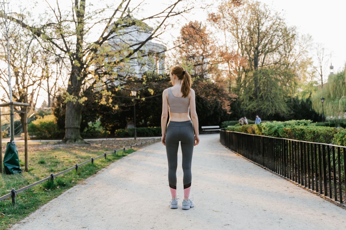 Woman in athletic wear walking along a sunlit park path lined with trees in the early morning