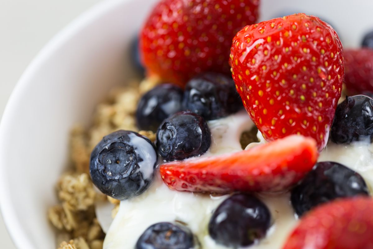 A bowl of thick Greek yogurt topped with fresh berries and seeds beside a spoon on a light surface